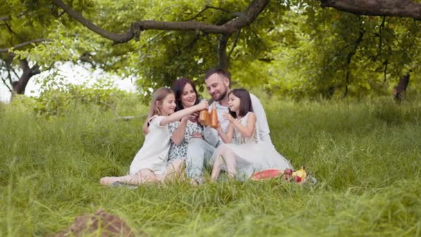 Genitori sorridenti e le loro belle figlie sedute su morbidi plaid all'aperto e brindare con bottiglie di succo fresco. Famiglia al picnic. Concetto di piacere. Figlie tostate con bottiglie di succo fresco. — Video Stock