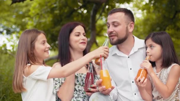 Padre sonriente, madre y dos hijas lindas que beben jugo fresco en el jardín verde mientras hacen un picnic de verano. Concepto de familia, ocio y relajación. Padres con dos hijas bebiendo jugo — Vídeo de stock