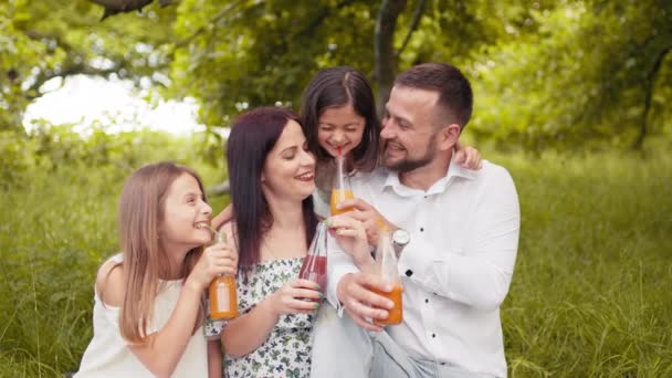 Glad ung familj med två barn som dricker färsk juice medan de sitter på filt i grön trädgård. Leende föräldrar och deras vackra döttrar sitter på mjuka rutiga utomhus och skålar med flaskor — Stockvideo