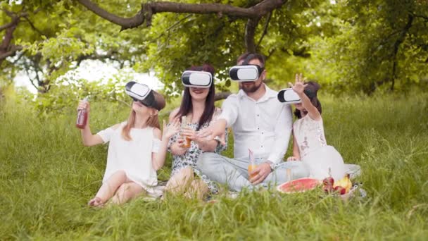 Parents and their two daughters wearing VR headset during picnic at summer garden. Young family having fun with innovative gadgets outdoors. Modern lifestyles. Parents with two kids at outdoors picnic — ストック動画