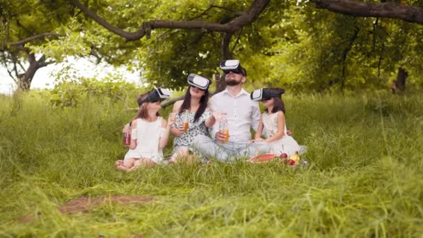 Parents and their two daughters wearing VR headset during picnic at summer garden. Young family having fun with innovative gadgets outdoors. Modern lifestyles. Parents with two kids using VR — ストック動画