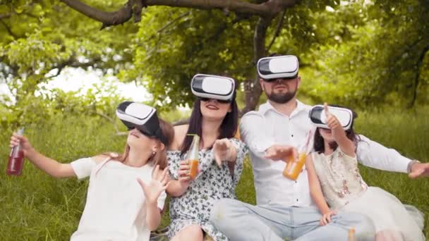 Young caucasian family having fun in virtual glasses on fresh air. Happy parents with two daughters sitting on soft blanket and playing games. Leisure time and entertainment concept. Parents using VR — ストック動画