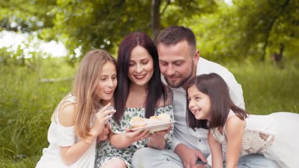 Mladí rodiče se dvěma dětmi ochutnávají vynikající dortíky, zatímco spolu sedí na měkké přikrývce venku. Pojem rodina, relaxace a požitek. Šťastná rodina jíst chutné cupcakes na piknik — Stock video