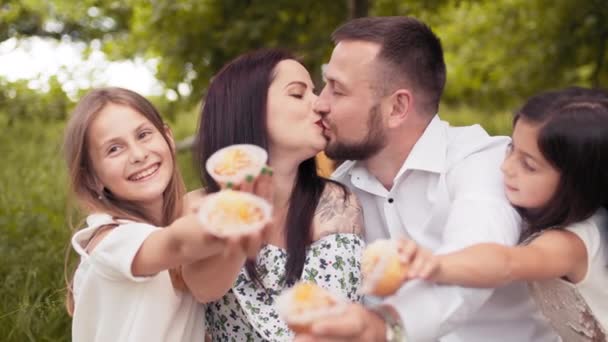 Hombre guapo y encantadora mujer besándose mientras sus dos hijas lindas sosteniendo cupcakes en las manos sonriendo en la cámara. Felicidad dentro de la familia caucásica. Hermanas comiendo cupcakes mientras los padres se besan — Vídeo de stock