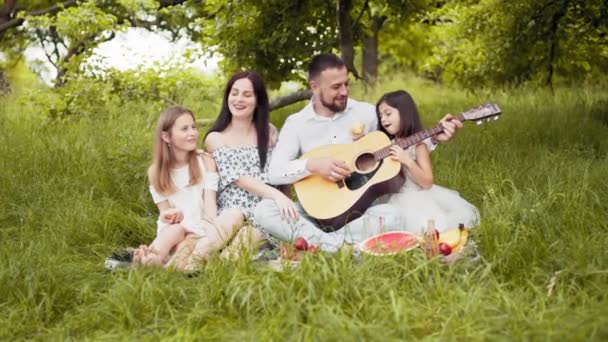 Handsome man playing guitar for his charming wife and two cute daughters. Happy caucasian family sitting on soft blanket at garden and singing songs. Parents with two kids playing guitar on fresh air — ストック動画