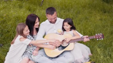 Handsome man playing guitar for his charming wife and two cute daughters. Happy caucasian family sitting on soft blanket at garden and singing songs. Parents and kids spending leisure time together