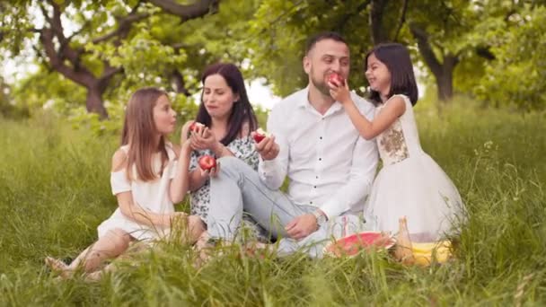 Retrato de pais jovens positivos com duas filhas pequenas degustando maçãs vermelhas no jardim verde. Tempo de lazer com a família. Tempo de verão e humor alegre. Pais com crianças comendo maçãs vermelhas no piquenique — Vídeo de Stock