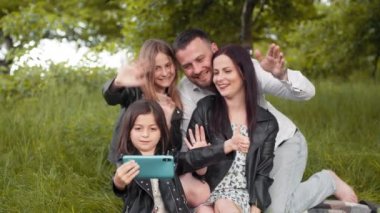 Positive family sitting together on soft plaid utdoors and taking selfie on modern smartphone. Young parents with two kids saving memories about happy summer picnic time. Cheerful parents with kids