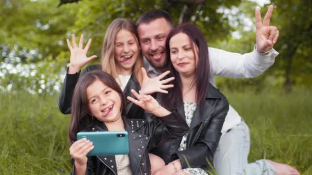 Familia positiva sentados juntos en tartán suave al aire libre y tomar selfie en el teléfono inteligente moderno. Padres jóvenes con dos hijos guardando recuerdos. Concepto de personas, ocio y tecnología. — Vídeo de stock