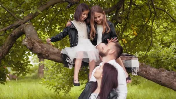 Bel marito che abbraccia la moglie incinta mentre le loro due figlie sono sedute su un ramo d'albero. Famiglia caucasica trascorrere del tempo insieme al verde giardino estivo. Famiglia in attesa posa in giardino — Video Stock