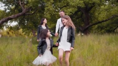Ebeveynleri el ele tutuşup arka planda yürürken iki neşeli kız kardeş birlikte yeşil bahçede oynuyorlar. Dört kişilik mutlu bir aile temiz havanın tadını çıkarıyor. İki çocuklu aile
