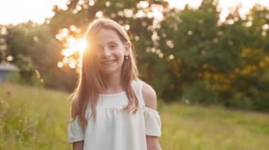 Pretty young girl wearing stylish white dress posing at green garden. Beautiful summer sunrise on background. Concept of childhood and carefree. Cute girl smiling and looking at camera at garden