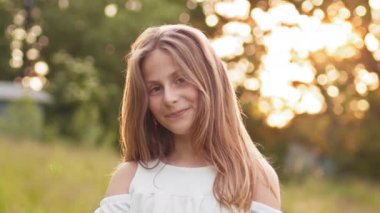 Attractive young blonde teen girl walking in summer sunset park, looks at camera with alluring look. Portrait of a young woman outdoors. Happy little girl, Children in park