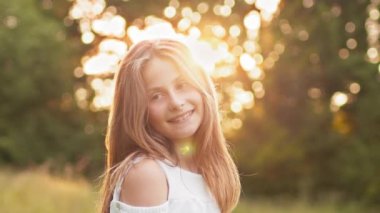 Pretty blond teenage girl with wavy hair smiling on camera looking cheerful. Positive emotions. Attractive young blonde teen girl walking in summer sunset park, looks at camera with alluring look.