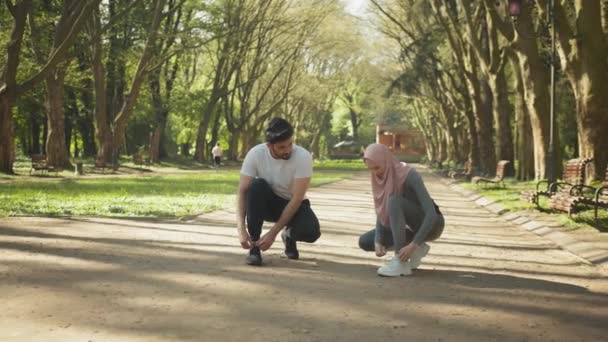 Arabski mężczyzna i kobieta wiążą sznurowadła na tenisówkach sportowych. Muzułmanin i kobieta w hidżabie wiążą sznurowadła na tenisówkach sportowych przed porannym bieganiem po zielonym parku. Młode aktywne szkolenie rodzinne na świeżym powietrzu. — Wideo stockowe
