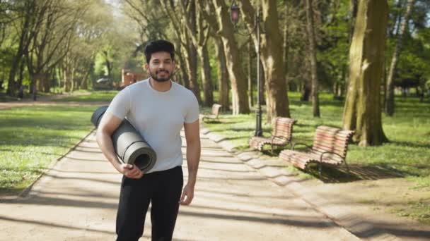Портрет усміхненого арабського чоловіка в спортивному одязі після тренування в парку. Молодий активний хлопець позує на відкритому повітрі з йога мат в руках. Усміхнений арабський чоловік у спортивному одязі в парку — стокове відео
