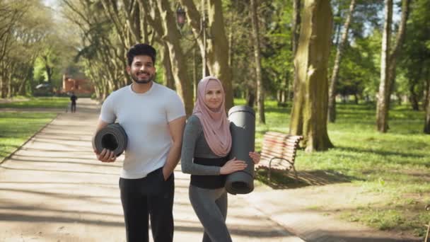 Przedni widok uśmiechniętego muzułmanina i czarującej kobiety w hidżabie stojącej razem w zielonym parku i trzymającej maty do jogi. Szczęśliwa rodzina prowadzi aktywny i zdrowy tryb życia. Arabska para w odzieży sportowej — Wideo stockowe