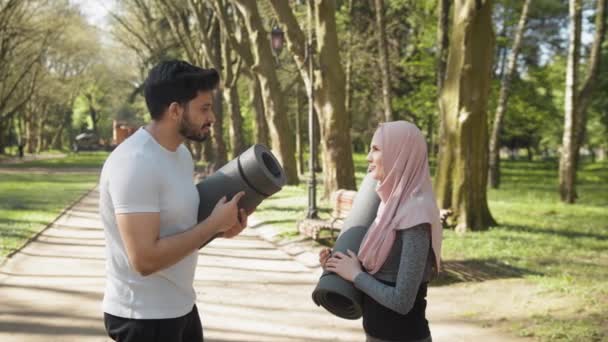 Arabska para w stroju sportowym spogląda na siebie stojąc w zielonym parku miejskim i trzymając maty do jogi. Szczęśliwa rodzina spędza wolny czas na treningach na świeżym powietrzu. Uśmiechnięty muzułmanin mężczyzna i kobieta — Wideo stockowe
