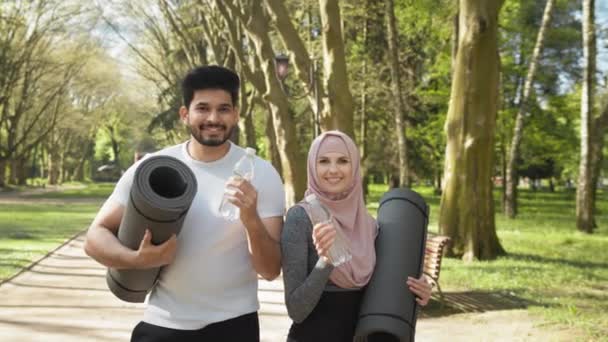Szczęśliwa muzułmanka i mężczyzna wznoszący toast butelką świeżej wody po treningu w zielonym parku. Młoda para w aktywnych ubraniach trzymająca maty do jogi podczas odświeżania na świeżym powietrzu. Rodzina arabska z butelką wody — Wideo stockowe