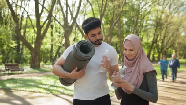 Młoda kobieta w odzieży sportowej i hidżab za pomocą nowoczesnego smartfona, a przystojny muzułmanin pije wodę i trzyma matę do jogi. Szczęśliwa para relaksuje się po treningu na świeżym powietrzu. Arabski mężczyzna i kobieta — Wideo stockowe