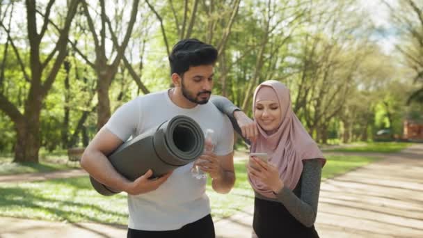 Schöner arabischer Typ mit Yogamatte und Wasserflasche, der seine charmante Frau im Hijab mit dem Handy anschaut. Junge Familie in Aktivkleidung steht am grünen Morgenpark. Sportliche Frau nutzt Handy — Stockvideo