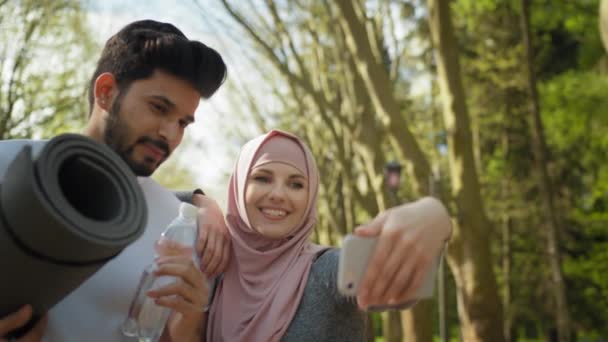 Vrolijke moslim man en vrouw in hijab nemen zelfportret op smartphone terwijl ze in het stadspark staan. Jong actief stel in sportkleding die pauze neemt tijdens de training. Moslim paar in sport kleding — Stockvideo