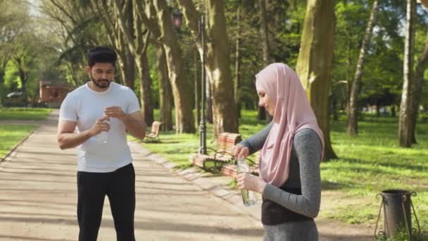 Piękna kobieta w strojach sportowych i hidżab trzymając butelkę wody stojąc w zielonym parku. Rozmyte tło przystojnego muzułmanina orzeźwiającego i relaksującego po bieganiu. Para w sportowej odzieży pozowała — Wideo stockowe