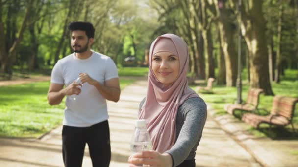 Csinos nő hidzsábban, üveggel a kezében, miközben a muszlim fickó félrenéz. Fiatal család aktív ruházat felújítása vízegyensúly után szabadtéri edzés. Muszlim pár. — Stock videók
