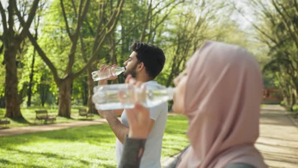 市内公園に立っている間、ボトルから新鮮な水を飲むアクティブな姿でイスラム教徒の女性と男性の側面図。若いカップルは屋外での朝の運動中に休憩。スポーツ衣装のイスラム教徒のカップル — ストック動画