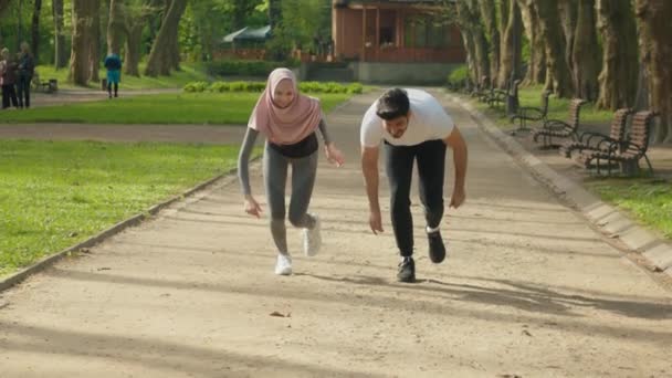 朝の公園で一緒に走っているスポーツ服の肯定的な若者。野外で積極的にヒジャーブ訓練でハンサムなイスラム教徒の夫と魅力的な妻。幸せなアラビア語カップルでアクティブな摩耗ジョギング — ストック動画