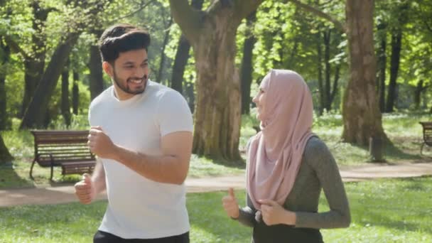 緑の公園では、朝の時間帯にアラビア人のカップルがジョギングをしています。若い男性と女性が一緒に過ごす時間を楽しんでいます。屋外で一般的なワークアウト。公園で一緒に走る幸せなアラビア人のカップル — ストック動画