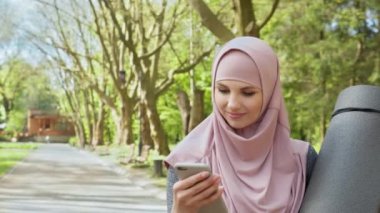 Açık hava egzersizi sırasında modern cep telefonu kullanan tesettürlü ve spor kıyafetli gülümseyen bir kadının ön görüntüsü. Aktif yaşam tarzı konsepti. Tesettürlü kadın yoga minderi tutuyor ve akıllı telefon kullanıyor.