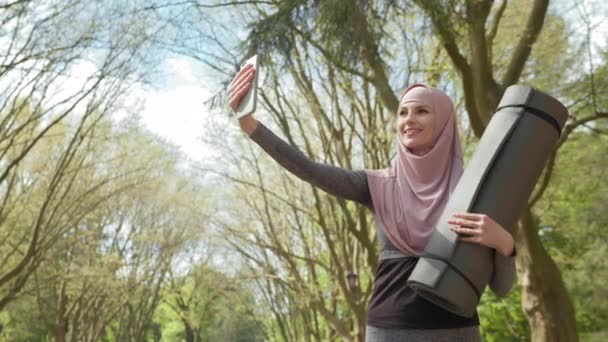 Szczęśliwa muzułmanka w stroju sportowym i hidżab robiąca selfie na nowoczesnym smartfonie stojąc w zielonym parku z matą do jogi w ręku. Koncepcja p ludzi, technologii i działalności. Muzułmanka selfie — Wideo stockowe