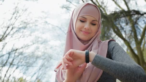 Attractive muslim woman in sport clothes and hijab checking pulse on smart watch. Female athlete with bottle of water relaxing after morning run. Woman in hijab checking jogging distance on watch — Stock Video