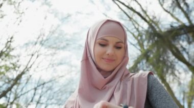 Tesettürlü hoş bir kadın yürüyüş mesafesini kontrol etmek için akıllı iz sürücüye bakıyor. Yeşil Park 'ta spor kıyafetli genç bir bayan atlet. Çalışan Çalıştırma Çalışmasında Nabız Kontrol Ediyor