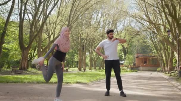 夏の公園でスポーツ服やヒジャーブストレッチ足でアクティブな女性。若いアラビア人の男が背景に立ってスマートウォッチを使用しています。屋外スポーツ活動。腕時計で男を暖める女 — ストック動画