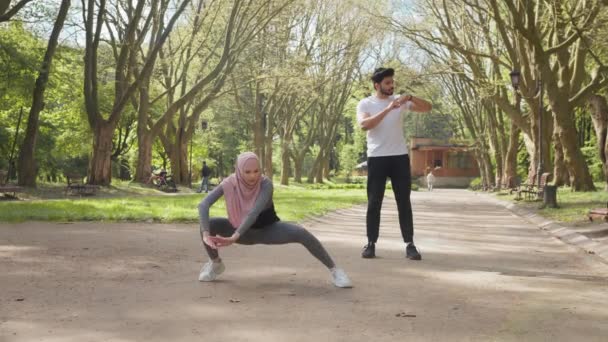 Μια γυμνασμένη γυναίκα με χιτζάμπ και αθλητικό ντύσιμο να ζεσταίνεται στο πάρκο πριν το πρωινό τρέξιμο. Όμορφος μουσουλμάνος που στέκεται στο παρασκήνιο και ελέγχει τον παλμό στο έξυπνο ρολόι. Γυναίκα που τεντώνει πόδια — Αρχείο Βίντεο
