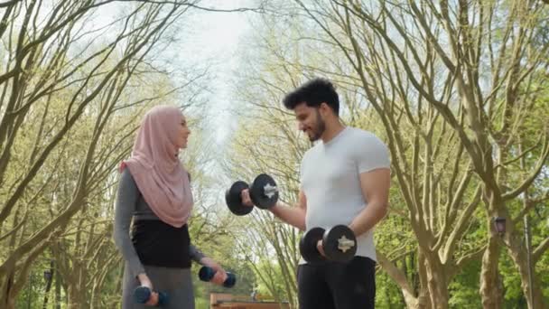 Jovem mulher em hijab e belo homem muçulmano de pé juntos no parque verde e braços de treinamento usando halteres. Casal feliz com treino de equipamento desportivo ao ar livre. Casal árabe usando halteres — Vídeo de Stock