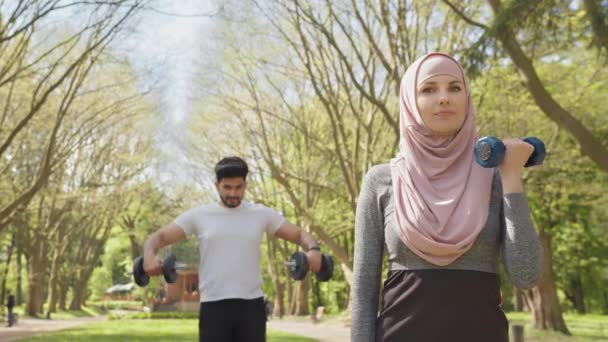 머리좋은 아라비안 호스 남자가 배경 훈련을 받는 동안 히잡의 건강 한 사타구니 여자와 함께 무기를 위한 운동을 하고 있습니다. 사람, 스포츠, 그리고 건강에 대한 개념. 히잡을 입은 여자와 그 근육질 남자 — 비디오
