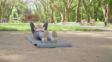 Tesettürlü, spor elbiseli, yoga minderinde oturan ve vücudu esneyen hoş bir kadın. Genç Müslüman kadın eğitimi sabah vakti açık havada. Müslüman kadın yaz parkında yoga minderi eğitimi alıyor.