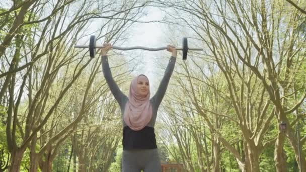 Sportig kvinna i aktiva kläder och hijab håller tung skivstång över huvudet när du står i parken. Glad muslimsk kvinnlig träning på fritiden utomhus. Kvinna i hijab håller skivstång över huvudet — Stockvideo