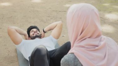 Hintli kaslı adam ayaklarını tutan tesettürlü kadının yardımıyla karın kaslarını yoga minderinde çalıştırıyor. Aktif genç çift temiz havada birlikte antrenman yapıyor. Hintli adam karın kası çalıştırıyor.