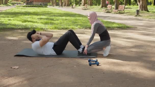 Glad aktiv man i sportkläder sitter på yoga man och gör magövningar. Baksidan av kvinnan i hijab håller manliga fötter under träningen. Begreppet hälsa och kroppsvård. Man gör övning för abs — Stockvideo