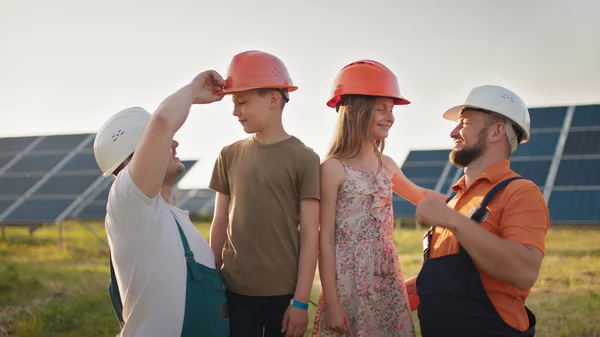 Portrett av to fedre og to barn i beskyttende hjelmer ved et solkraftverk, som alle peker fingre mot kameraet og smiler. Foreldrene satte en hjelm på barnas hoder ved et solkraftverk. – stockfoto