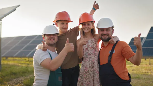 Retrato de dois pais e duas crianças em capacetes de proteção em uma usina de energia solar, todos apontando os dedos para a câmera e sorrindo. Os pais puseram um capacete nas cabeças das crianças numa central solar. — Fotografia de Stock