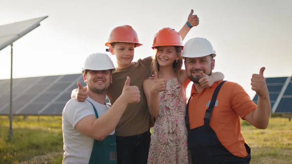 Retrato de dois pais e duas crianças em capacetes de proteção em uma usina de energia solar, todos apontando os dedos para a câmera e sorrindo. Os pais puseram um capacete nas cabeças das crianças numa central solar. — Fotografia de Stock