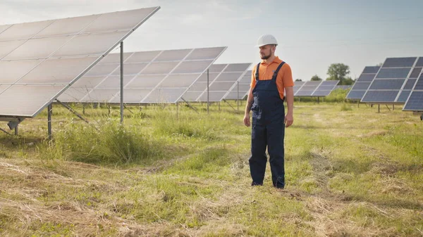 Ammattimies teknikko kypärä kävelee uuden ekologisen aurinkorakenteen ulkona. Aurinkopaneelien tila. Käsite sähkö, ekologia, teknologiat. Kameraa etsivän insinöörin muotokuva — kuvapankkivalokuva