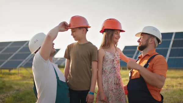 Portrett av to fedre og to barn i beskyttende hjelmer ved et solkraftverk, som alle peker fingre mot kameraet og smiler. Foreldrene satte en hjelm på barnas hoder ved et solkraftverk. stockfoto