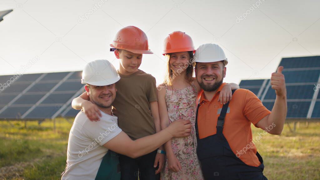 Portrait of two dads and two kids in protective helmets at a solar power plant, all pointing fingers up at the camera and smiling. Parents put a helmet on their childrens heads at a solar power plant