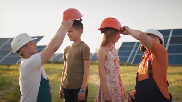 Çocuklar aileleri için çalışmaya geldiler. İki baba çocukları için kask takar ve iş yerinde sarılırlar. İş yerinde çocukları kameraya gülümseyen bir güneş enerjisi santrali çalışanı. Babam mutlu. — Stok video